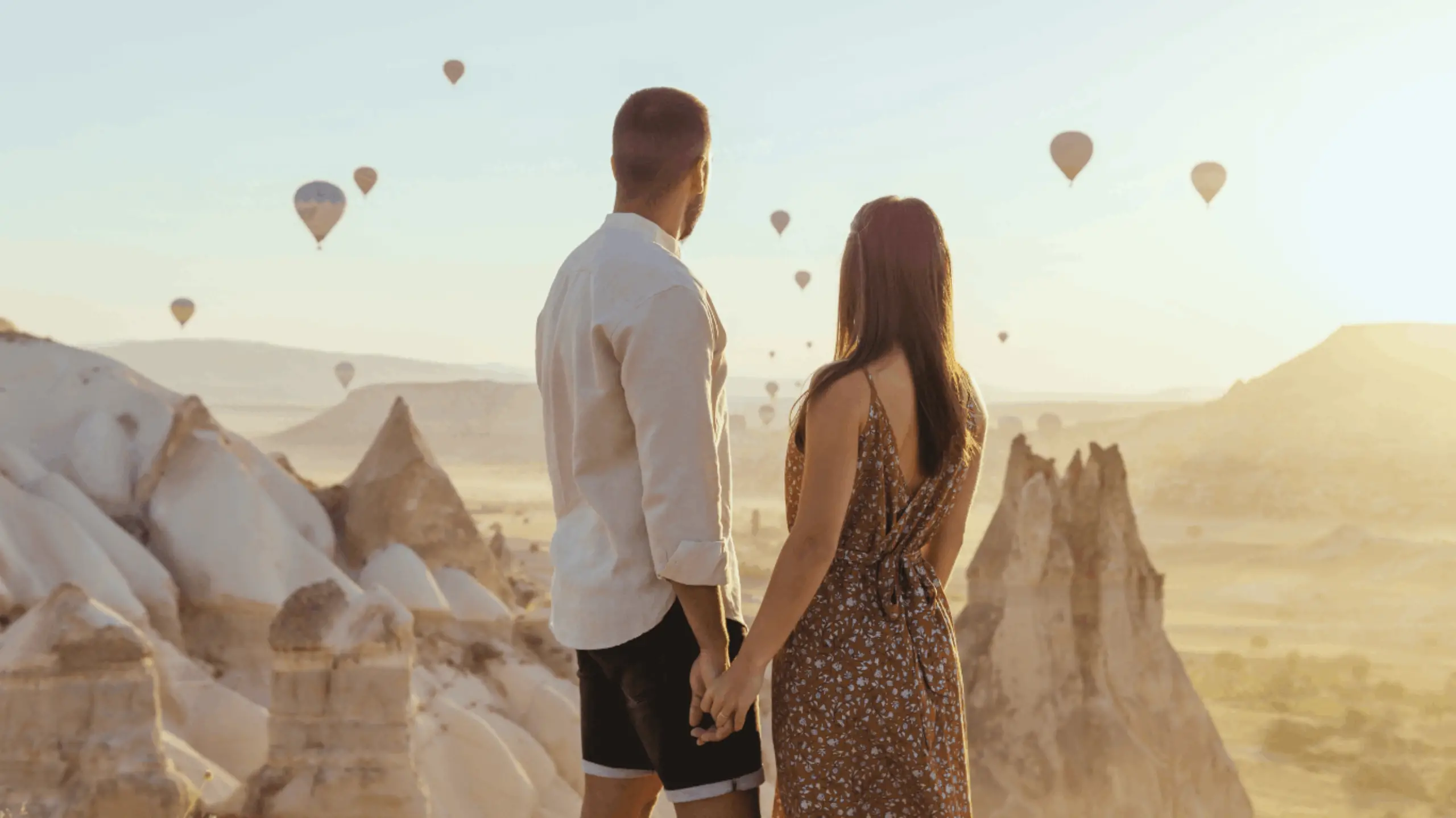 Foto de una pareja donde el hombre le está tocando la mano a una mujer y ambos están mirando al horizonte donde hay globos. Es una ilustración dentro de la página de Lovitmatch Making.